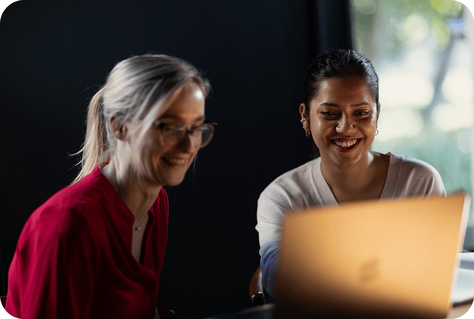 Two women smiling while discussing something on a laptop screen.