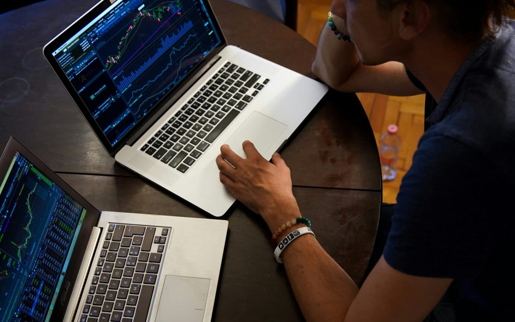 A person is sitting at a round table using two laptops, both displaying stock market charts and graphs. They are leaning on one arm, focused on the screens.